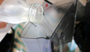 A hand pouring vinegar from a glass jar into the reservoir of a Keurig coffee maker.